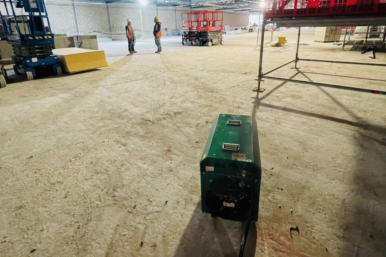 Electric Fan Heater 18KW in the foreground supplying heat to a large industrial warehouse with workers talking in the background and cherry pickers off to the side