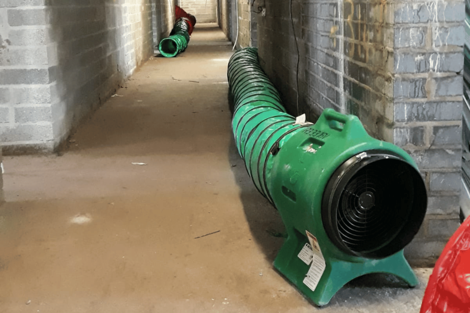 Climex Air Distribution Kit 300 in the foreground of a hallway with visibly damp bricks and flooring, receiving hot air to circulate and help dry the building