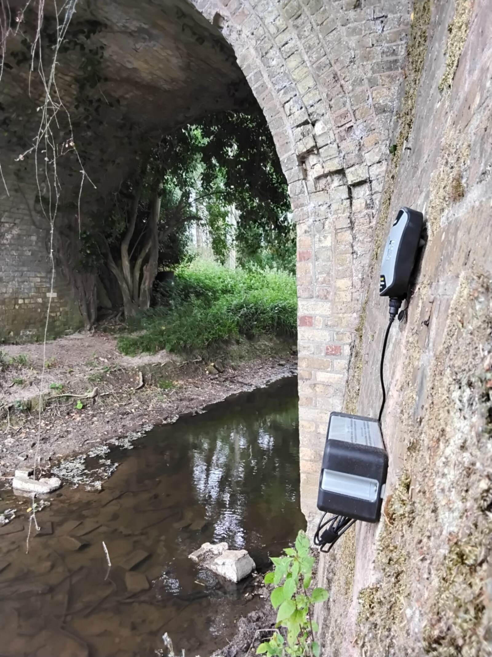 MONITEX OD-AA Vibration Monitor mounted on a wall under a bridge with a stream running beneath it