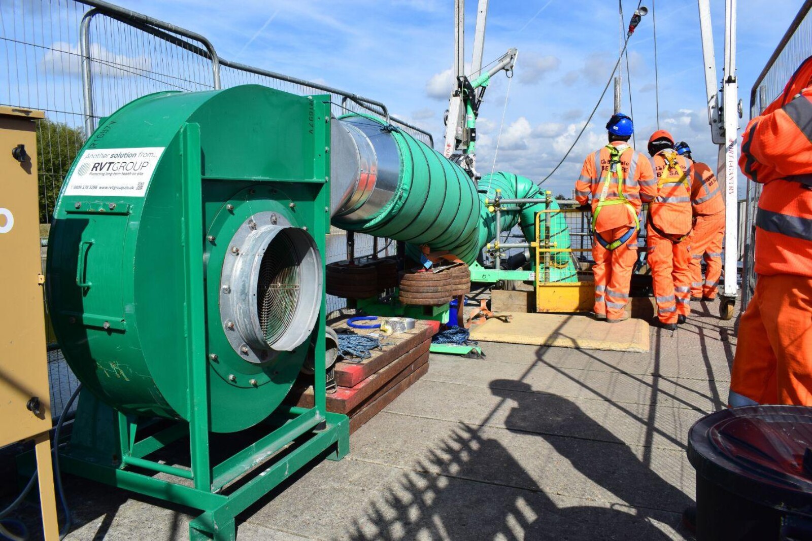 VENTEX Centrifugal Fan 450S with ducting entering a small hole on the platform nearby. Workers in orange overalls stand next to the hole with water in the background